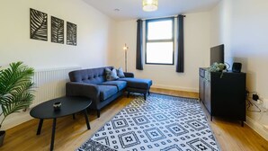 Un salon avec un canapé, une table basse, un tapis à motifs, une plante et un téléviseur posé sur un meuble.