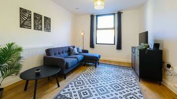 A living room with a sofa, a coffee table, a patterned rug, a plant, and a TV on a cabinet.