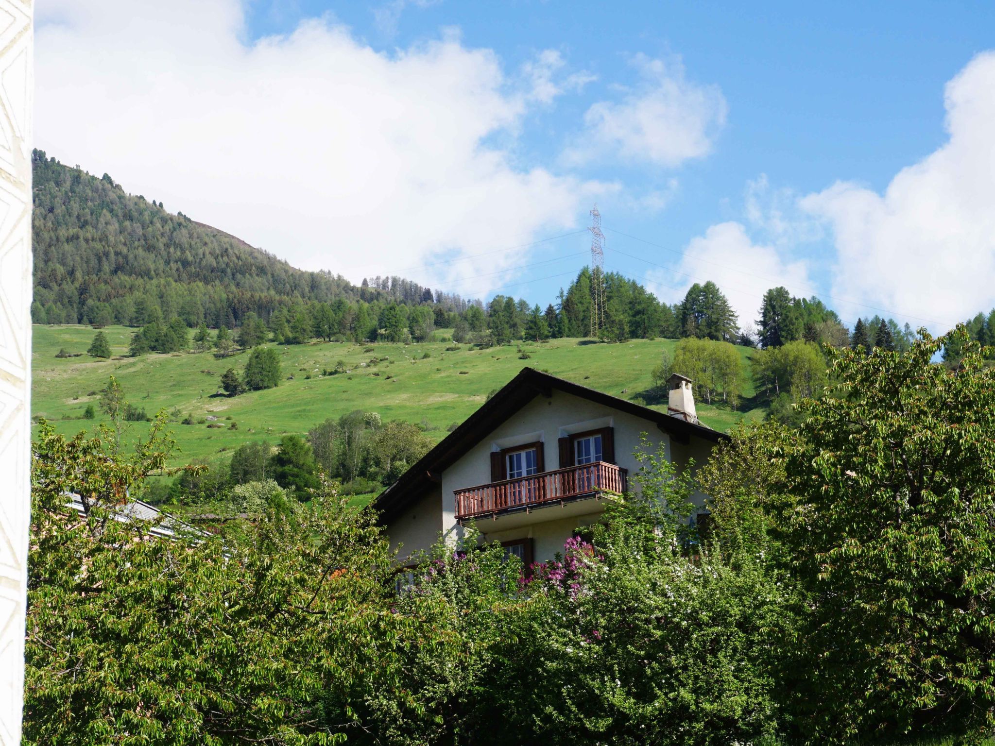 Ein zweistöckiges Haus mit Balkon, umgeben von üppiger Vegetation und einer hügeligen Landschaft.