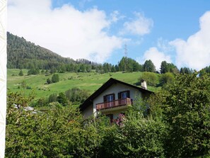 Een twee verdiepingen tellend huis met een balkon, omringd door weelderige begroeiing en een glooiend landschap.
