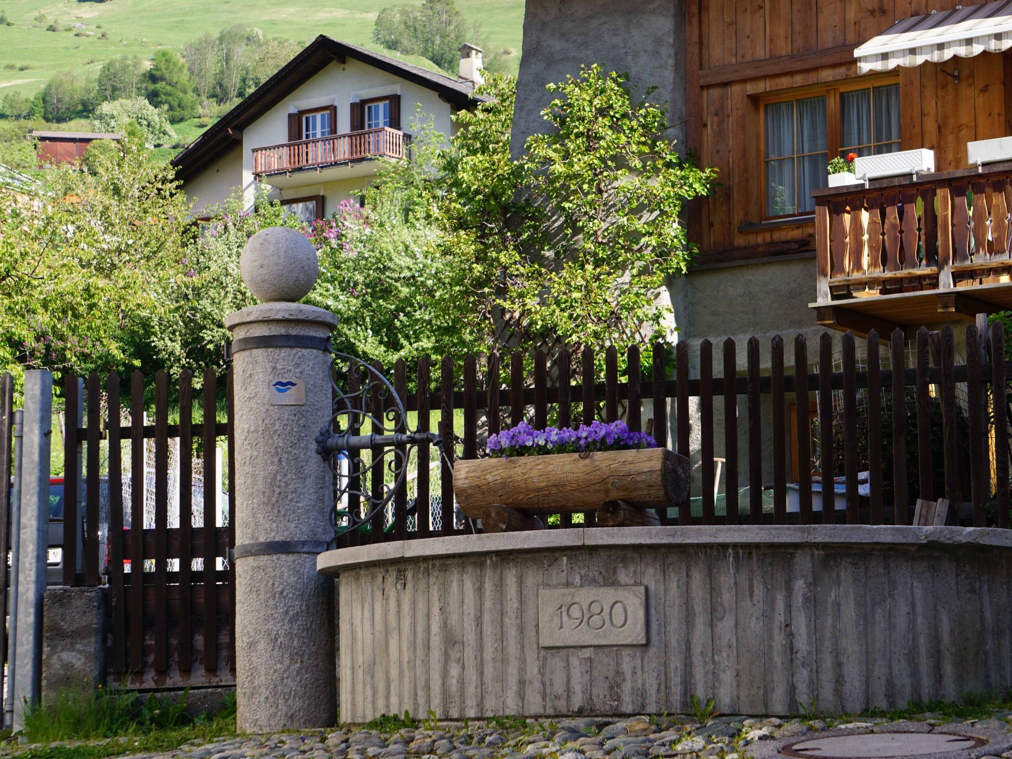 Eine steinerne Säule mit einer Gedenktafel, ein Holzzaun und ein Blumenbeet mit violetten Blumen.