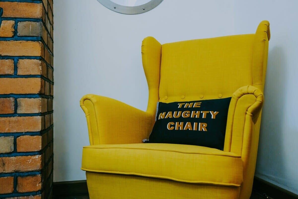 A yellow armchair with a pillow that reads 'THE NAUGHTY CHAIR' against a plain wall.