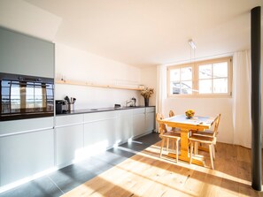 A modern kitchen with a dining area, featuring a wooden table and chairs, a built-in oven, and a window providing natural light. - Chasa Muntanella, (Ftan) by Interhome (Ftan)