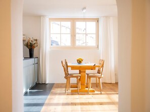 A dining area with a wooden table and chairs, a window with curtains, and a vase with flowers on a cabinet. - Chasa Muntanella, (Ftan) by Interhome (Ftan)