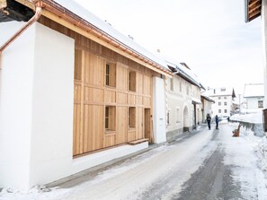 A snow-covered street with a building featuring large wooden windows and a white facade. - Chasa Muntanella, (Ftan) by Interhome (Ftan)