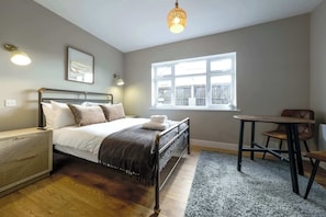 Ein Schlafzimmer mit Bett, Nachttisch, Lampe, Fenster und einem kleinen Tisch mit Stuhl.