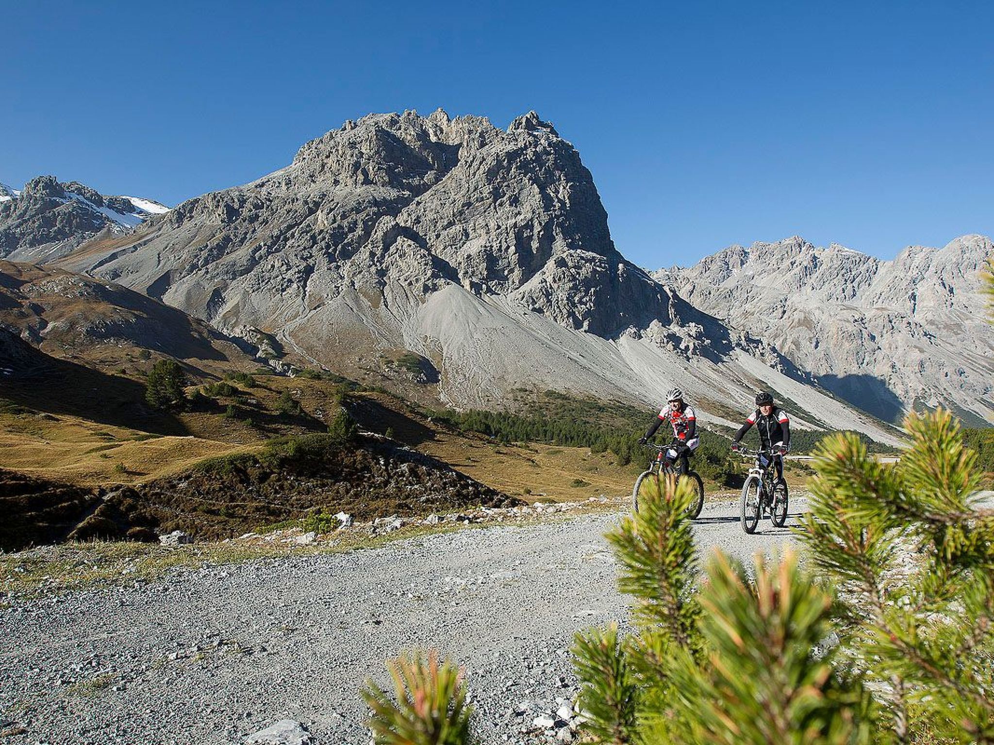 Un camino de grava con dos ciclistas, una montaña con cima y vegetación escasa.