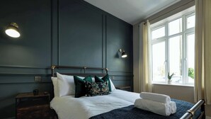 En pænt redt seng med hvide sengetæpper og et mørkeblåt tæppe, et sengebord med en lampe, og et vindue med gardiner.