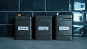 Tres recipientes negros etiquetados 'SUGAR', 'COFFEE' y 'TEA' sobre una superficie de azulejos.
