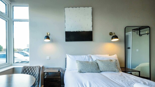 Una habitación de hotel con cama, mesita de noche, lámpara, silla y un ventanal grande.
