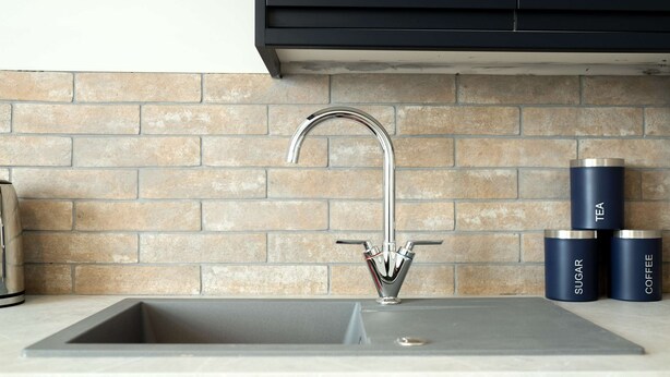 Una cocina con un grifo moderno, un respaldas de ladrillo y tres recipientes cilíndricos etiquetados 'TÉ', 'AZÚCAR' y 'CAFÉ'.