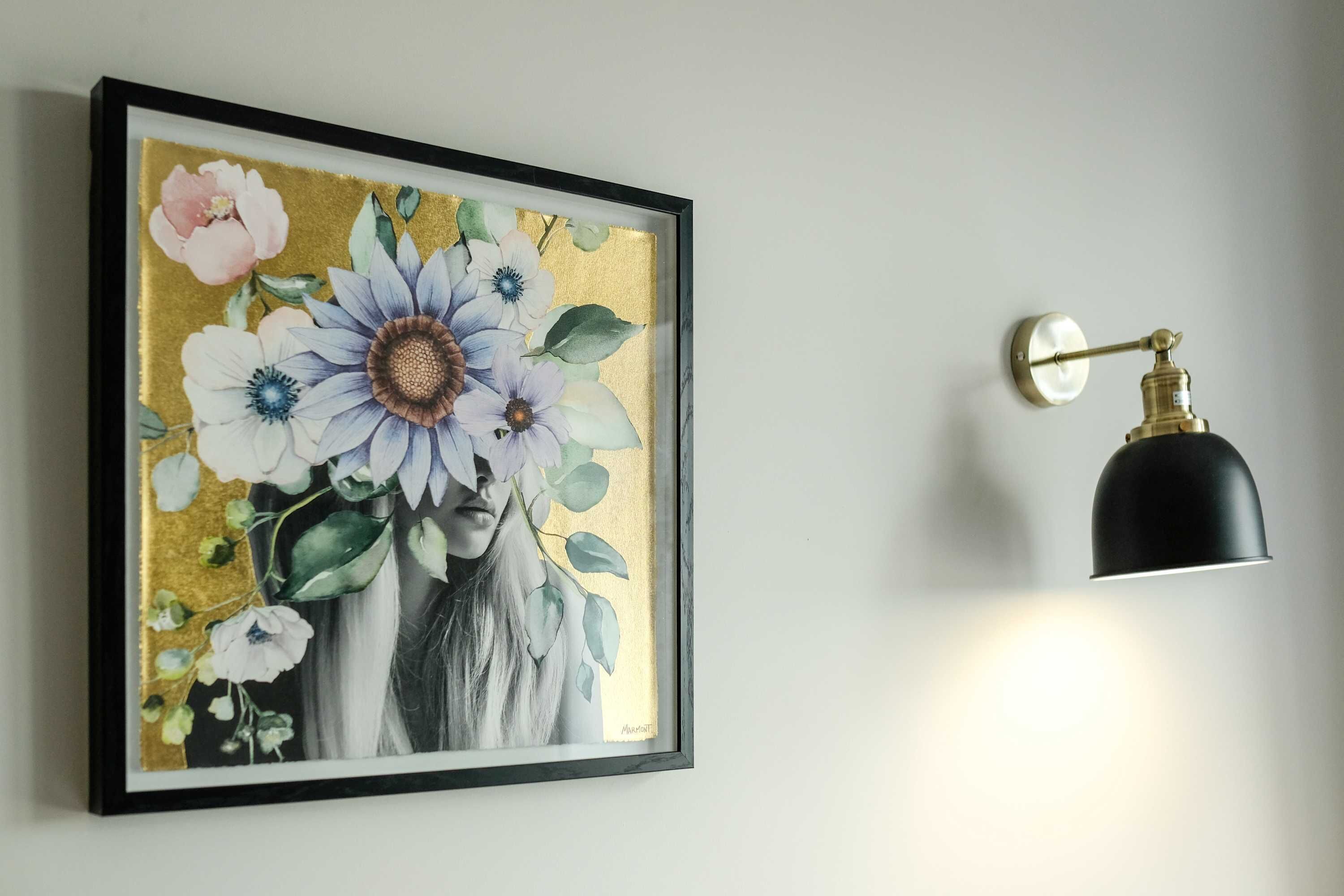 A framed floral artwork on a wall, a black wall-mounted light fixture, and a brass faucet.