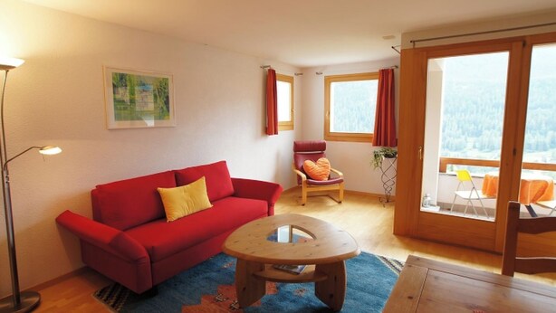 A cozy living room with a red sofa, a wooden coffee table, and a view of a forest through large windows. - TRAFÖGL, (Scuol) by Interhome (Scuol)