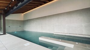 A modern indoor swimming pool with a minimalist design, featuring a clear water surface, a tiled wall, and a wooden ceiling.
