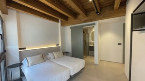 A modern hotel room with two beds, wooden ceiling beams, a small bedside table, and a bathroom visible through an open door.