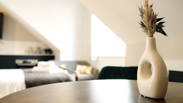 A modern living room with a round table, a vase with dried plants, and a view of a bedroom with a bed and a TV. - The Willow - Studio 14 (Patchway)