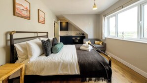 A bedroom with a bed, a bedside table, a window with a view, and a kitchenette in the background. - The Willow - Studio 14 (Patchway)