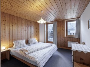 A wooden-paneled bedroom with a bed, bedside lamps, and a view of a snowy landscape through a window. - Plazzöl 111B, (Ftan) by Interhome (Ftan)