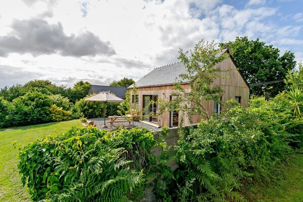 A rustic wooden house with a sloped roof, surrounded by lush greenery and a well-maintained lawn.