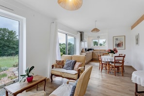 A spacious living area with a sofa, dining table, and chairs, a kitchen in the background, and large windows offering a view of the outdoors. - Sweet Escape in Brittany (Lanloup)