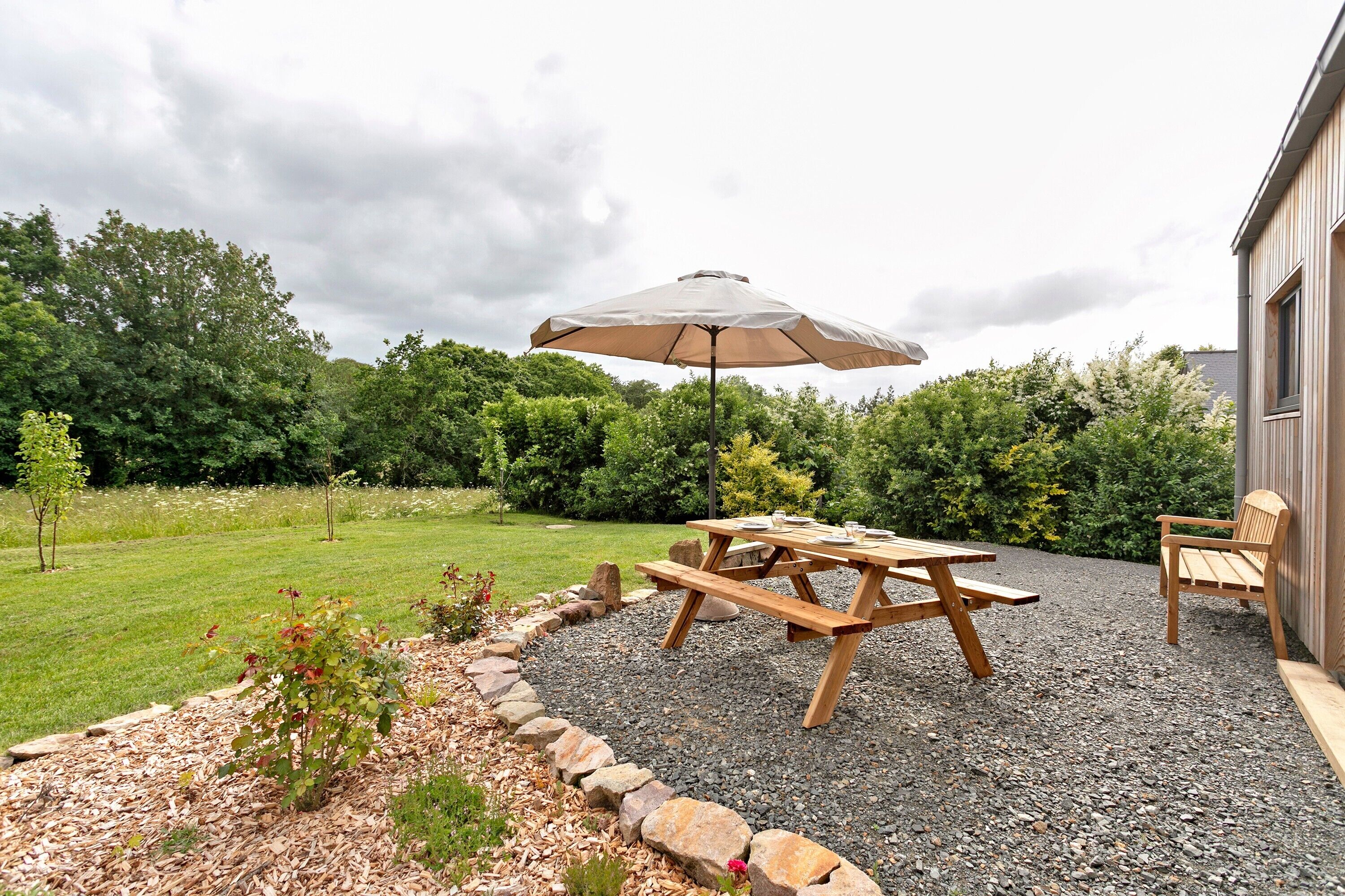 Een houten picknicktafel met een parasol, omringd door een grindpad met een stenen rand, en een grasveld op de achtergrond.