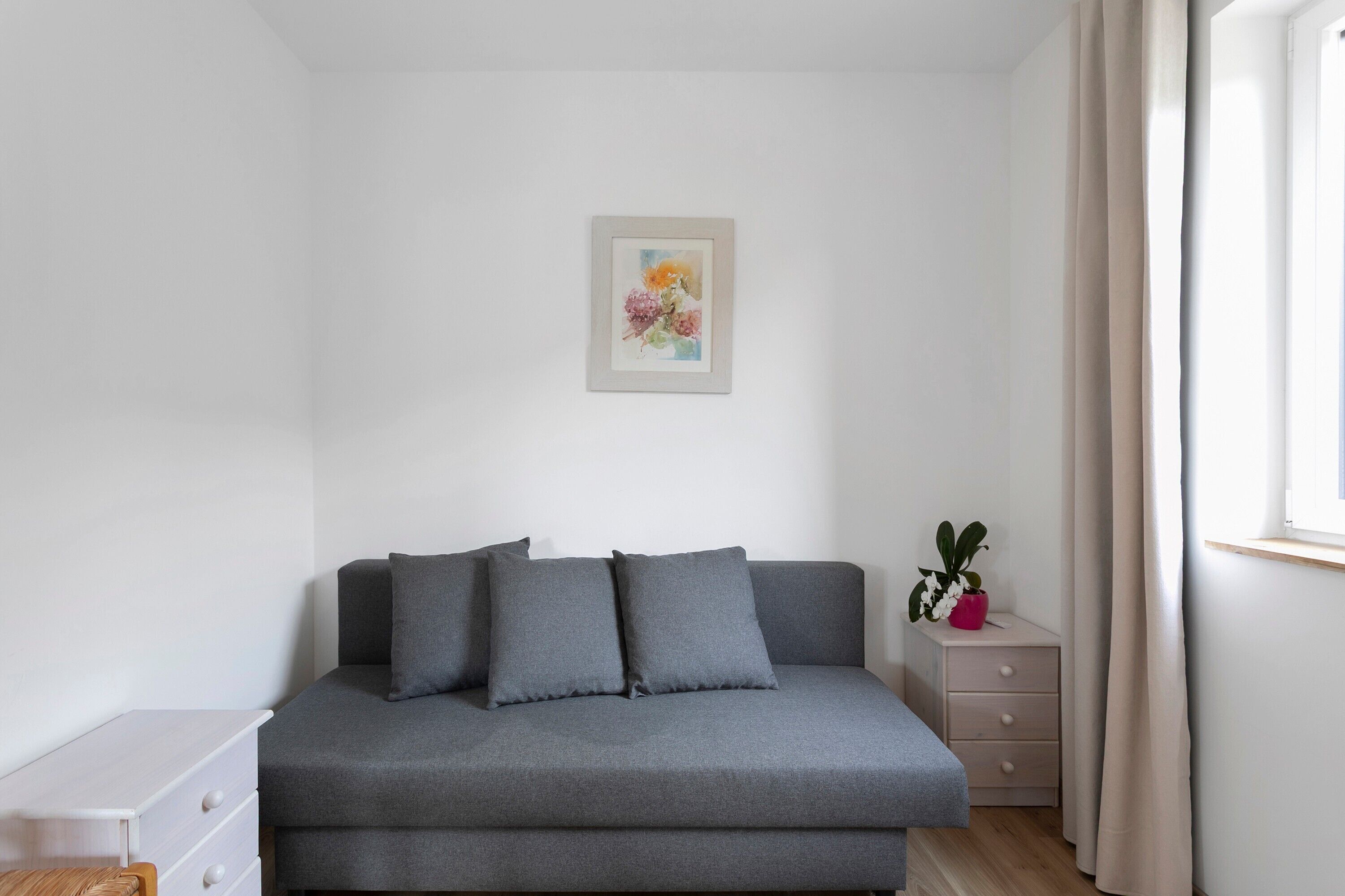Een minimalistisch ingerichte kamer met een grijze bank, een klein nachtkastje met een plant, een lijst met een foto aan de muur en een raam met gordijnen.
