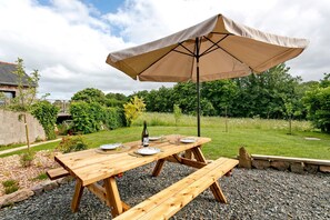 A wooden picnic table with an umbrella, surrounded by gravel and greenery.