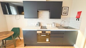 A compact kitchen with dark cabinets, a built-in oven, a sink, and a small dining area with a wooden table and a green chair.