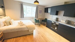 A modern hotel room with a bed, a dining table, a kitchen area, and a window with curtains.