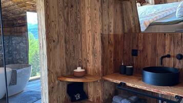 A rustic cabin interior with wooden walls, a stone fireplace, a bed with a view, and a small kitchen area.