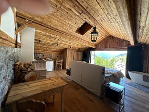 A rustic cabin interior with a wooden ceiling, a dining area, a kitchen, and a large window offering a mountain view.