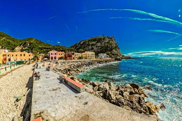 A coastal town with buildings along the beach, rocky shoreline, and a hill with a structure on top.