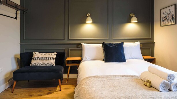 Une chambre d’hôtel comprenant un lit, deux tables de chevet, une chaise, une étagère fixée au mur et deux appliques murales.
