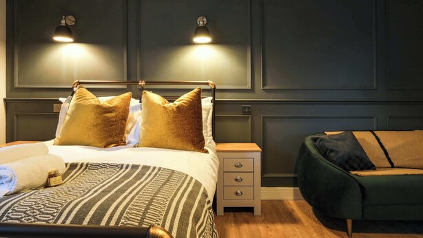 A bedroom with a bed, bedside tables, a nightstand, and a green sofa. - The Willow - Studio 27 (Patchway)