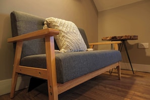 A wooden-framed sofa with a grey fabric cushion, a white knitted throw, and a small wooden side table with a rustic wood slab top. - The Willow - Studio 33 (Patchway)