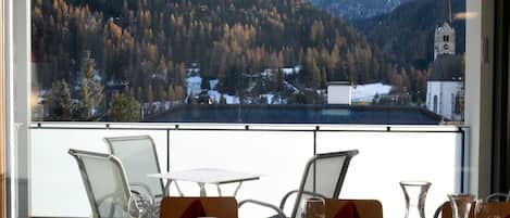 A dining area with a table set for two, overlooking a mountainous landscape with a church and a snow-covered village.