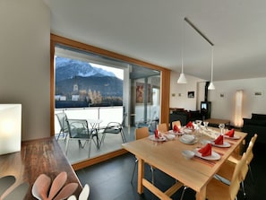 Un'area pranzo moderna con un tavolo di legno apparecchiato per quattro persone, ampie finestre con vista sulle montagne e un'accogliente zona salotto con camino.
