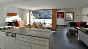 Une cuisine moderne avec un coin repas, de grandes fenêtres offrant une vue sur les arbres et les montagnes, et un salon avec un canapé et un tableau.