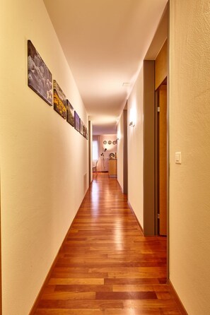 A hallway with wooden flooring, a door on the right, and framed pictures on the left wall.