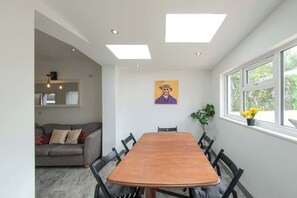 Un coin repas avec une table et des chaises en bois, un canapé, un tableau accroché au mur et une fenêtre donnant sur un paysage verdoyant.