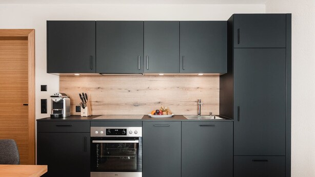 A modern kitchen with dark cabinetry, a built-in oven, a coffee machine, and a wooden backsplash. - Avero Lodge by MEIN SAMNAUN – MOUNTAIN HOMES, (Samnaun-Compatsch) by Interhome (Samnaun)