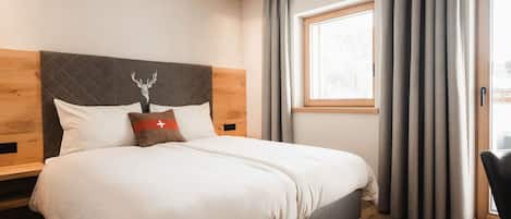 A modern bedroom with a large bed, a wooden headboard, a window with curtains, and a wooden floor.