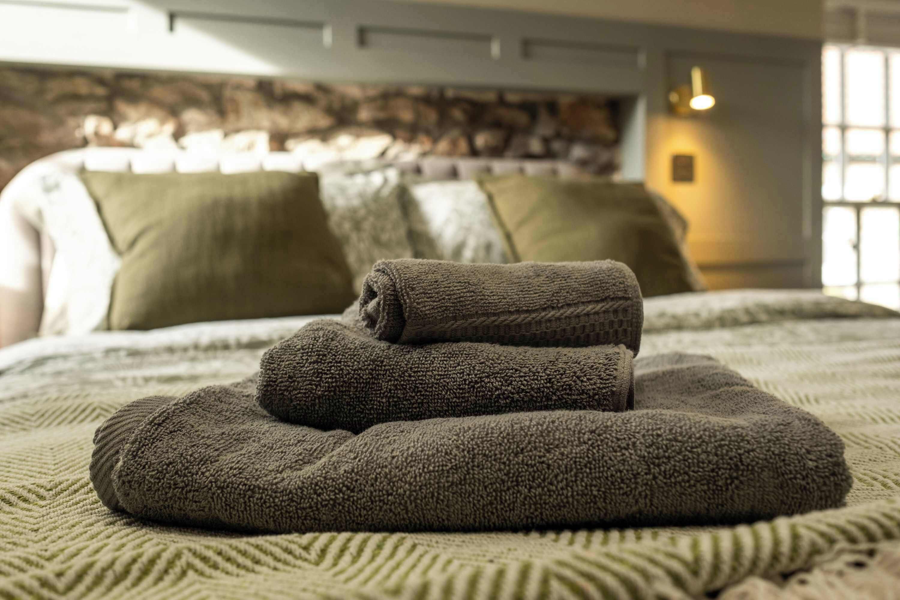 Uma cama bem arrumada com manta texturizada e almofadas, parede de pedra ao fundo e abajur de cabeceira.