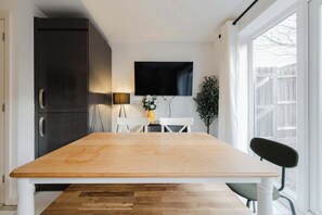 Eine Küche mit einem Holztisch, weißen Stühlen, einem schwarzen Kühlschrank, einem an der Wand montierten Fernseher und einem Fenster mit Blick auf einen Holzzaun.