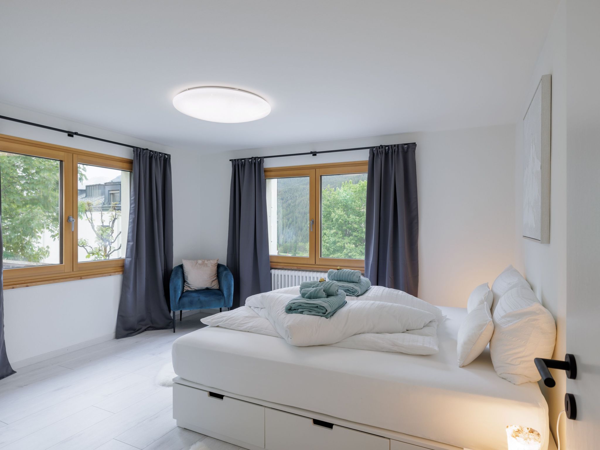 A modern bedroom with a bed, a chair, and two windows with curtains.