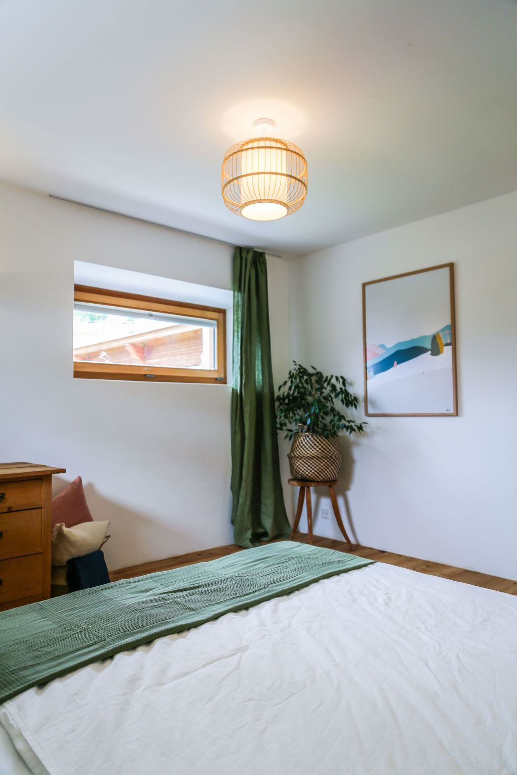 A bedroom with a bed, a window, a hanging light fixture, a plant, and a framed picture on the wall.