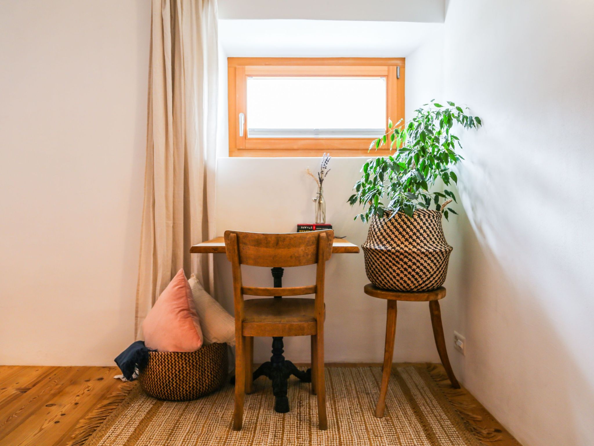 Um quarto pequeno com uma cadeira de madeira, uma mesinha, um vaso de planta e uma janela.