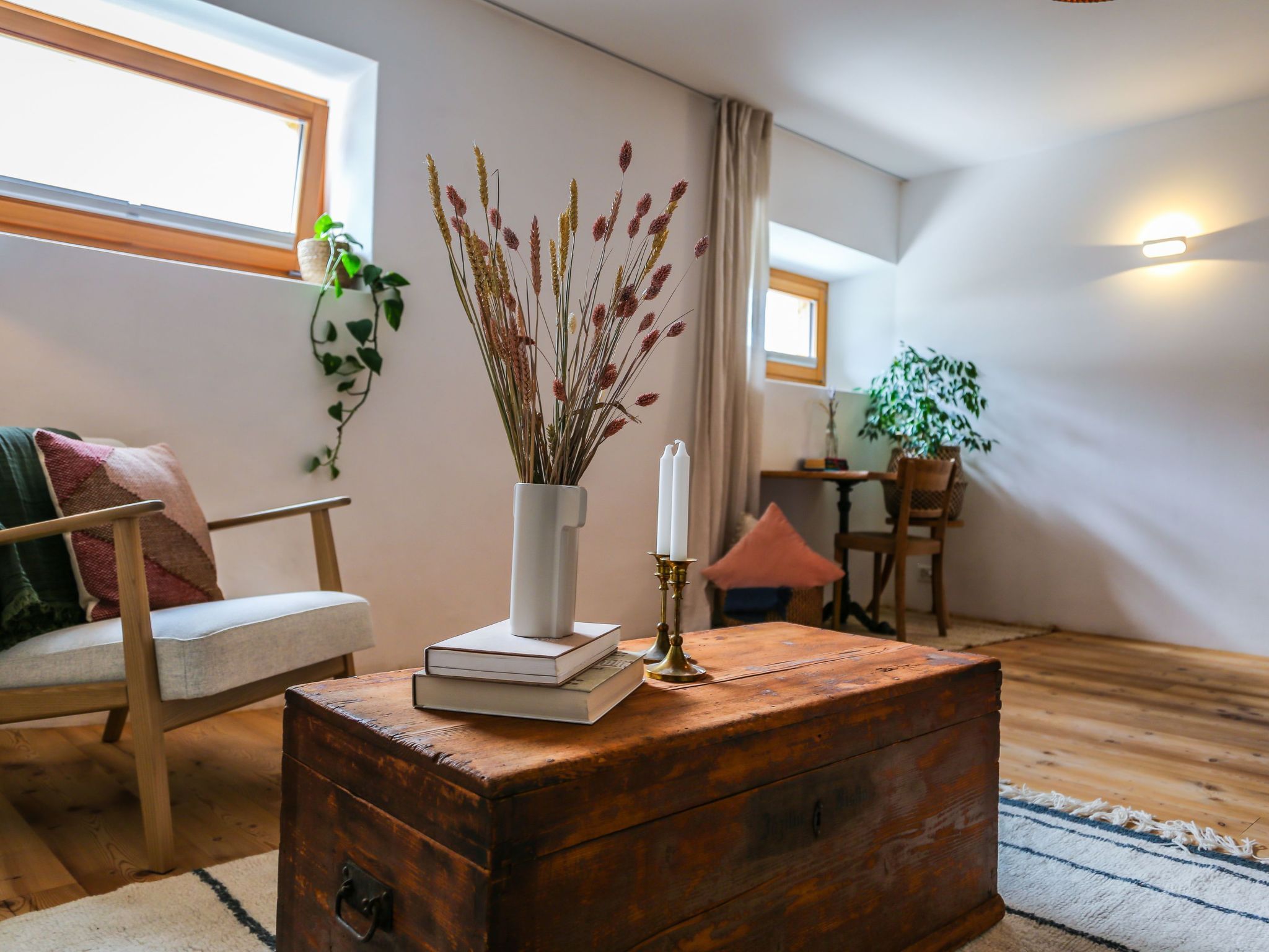 A cozy living room with a wooden chest, a white vase with dried plants, a bookshelf, and a window with curtains.