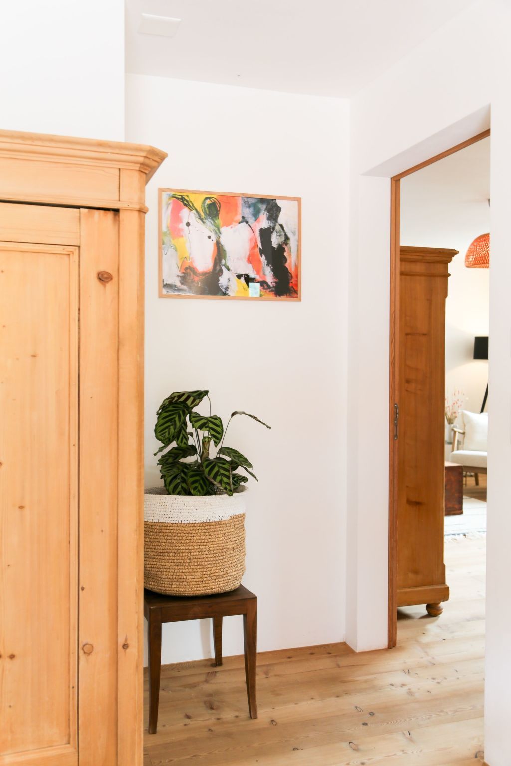 Un pasillo con un mueble de madera, una cesta tejida con una planta y un cuadro abstracto colorido en la pared.