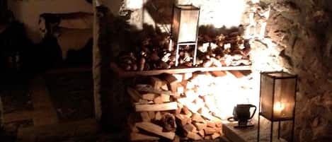 Interior rústico de piedra con chimenea, un estante con leña y un farol.